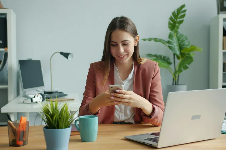 Jovem sorrindo enquanto responde mensagens no celular, sentada à mesa de trabalho
