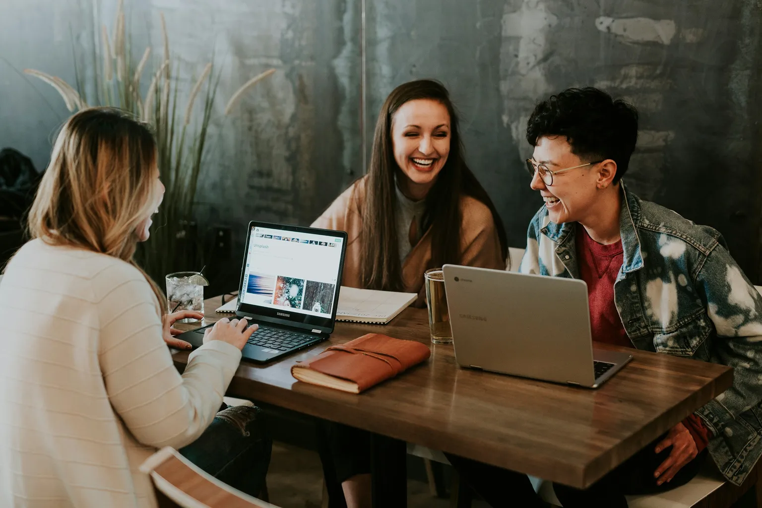 Três pessoas rindo juntas numa mesa de trabalho com notebooks